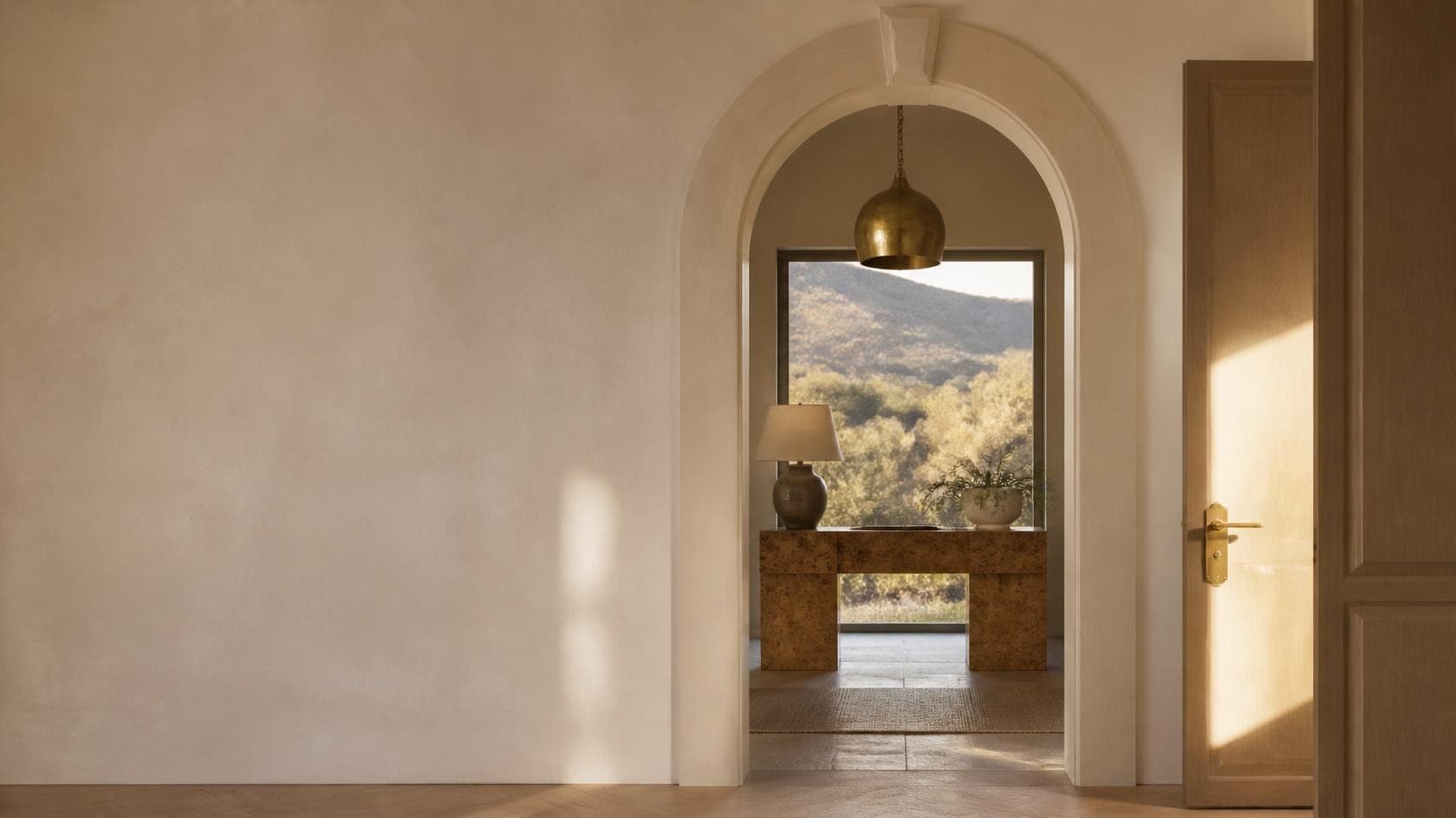 A plaster archway opening onto a brass pendant above a burl wood console, with a window onto oak-covered hills at sunset
