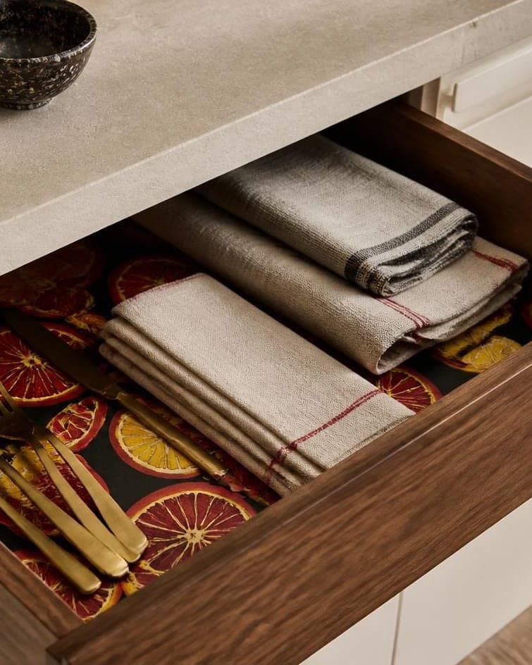 An open walnut drawer lined with a bold citrus-and-cherry paper, holding linen napkins and brass flatware